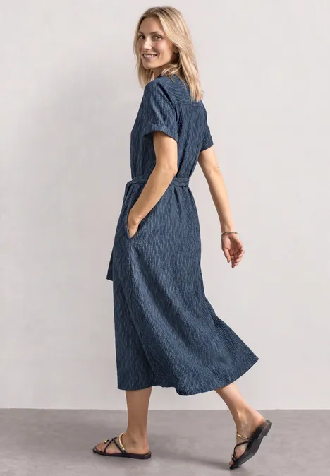 Midi-jurk met bowlingkraag darkest blue Vrouw in blauwe, midilengte jurk met zakken en zwarte sandalen tegen een neutrale achtergrond.