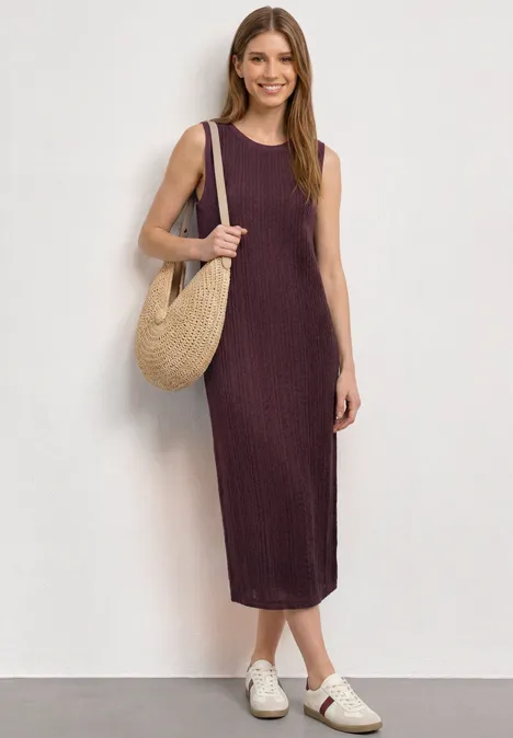 Midijurk van kant met ronde hals rustic rouge Vrouw in mouwloze, midilengte pruimkleurige jurk met rieten tas en witte sandalen tegen effen achtergrond.