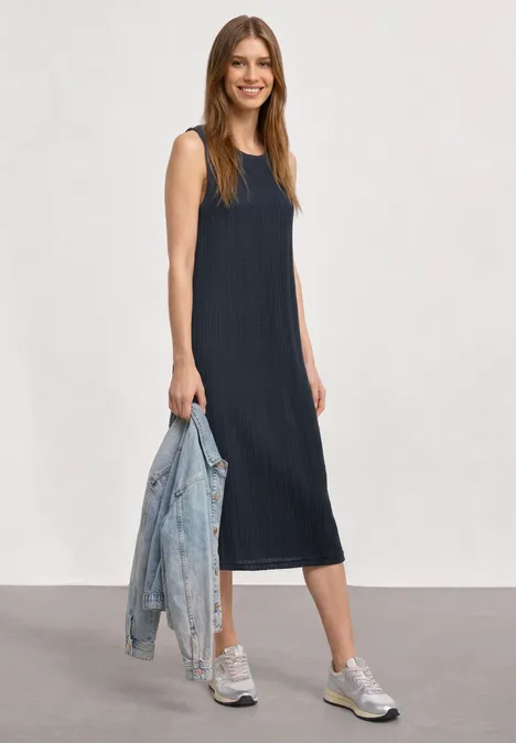 Midijurk van kant met ronde hals darkest blue Vrouw in mouwloze donkerblauwe midijurk met splitten, zilveren sneakers en houdt een spijkerjasje vast.