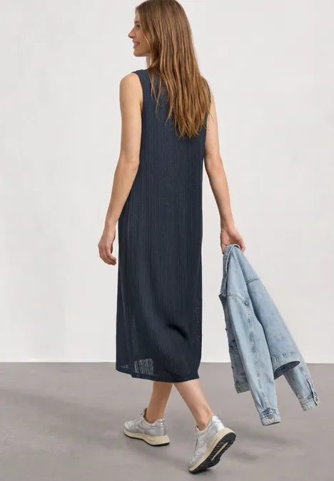 Midijurk van kant met ronde hals darkest blue Vrouw van achteren in donkerblauwe mouwloze midi-jurk en sneakers, draagt spijkerjasje in hand.