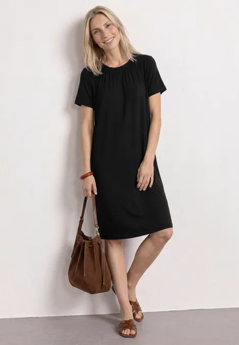 Knielange jersey jurk met ronde hals Black Vrouw in eenvoudige, knielange zwarte jurk met bruine handtas en sandalen, staand voor een witte achtergrond