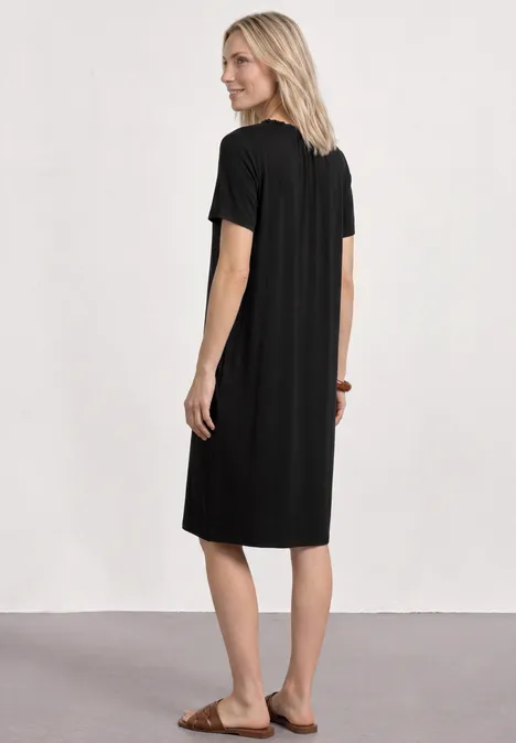 Knielange jersey jurk met ronde hals Black Vrouw met blond haar in een zwart midi-jurkje en sandalen, van achteren voor een witte muur.