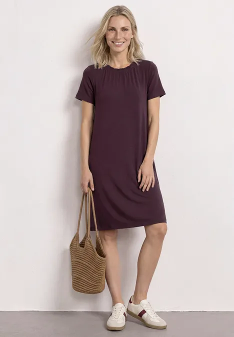 Knielange jersey jurk met ronde hals rustic rouge Een vrouw draagt een eenvoudig, knielang paars T-shirtjurkje, houdt een gevlochten strozak en witte sneakers.
