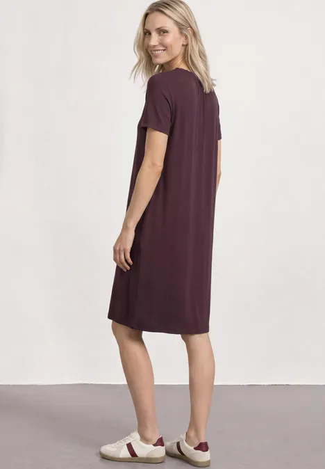 Knielange jersey jurk met ronde hals rustic rouge Een vrouw van opzij in een losvallende paarse T-shirtjurk en witte sportschoenen.