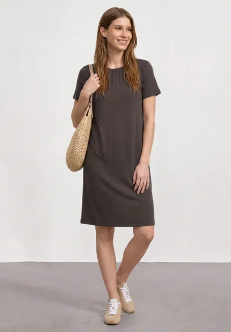 Knielange jersey jurk met ronde hals black coffee Jonge vrouw in knielange, donkergrijze T-shirtjurk met witte sneakers en gevlochten tas tegen neutrale achtergrond.