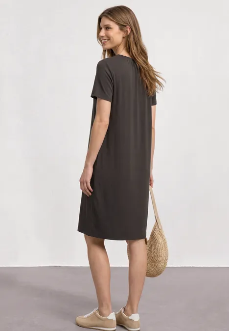 Knielange jersey jurk met ronde hals black coffee Vrouw van achter in losse, donkergrijze jurk tot op de knie, witte sneakers en strooien tas, lichte studioachtergrond.