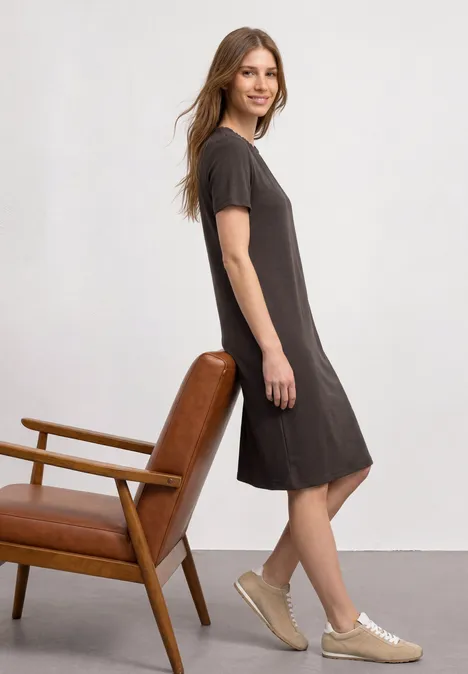 Knielange jersey jurk met ronde hals black coffee Vrouw in donkergrijze, knielange jurk en sneakers, schuin leunend tegen een houten leren fauteuil voor een witte muur.