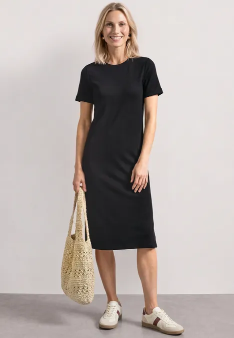 Midijurk met ribbelpatroon en ronde hals Black Vrouw in eenvoudige zwarte jurk, witte sneakers en nettas, staand voor een neutrale achtergrond.