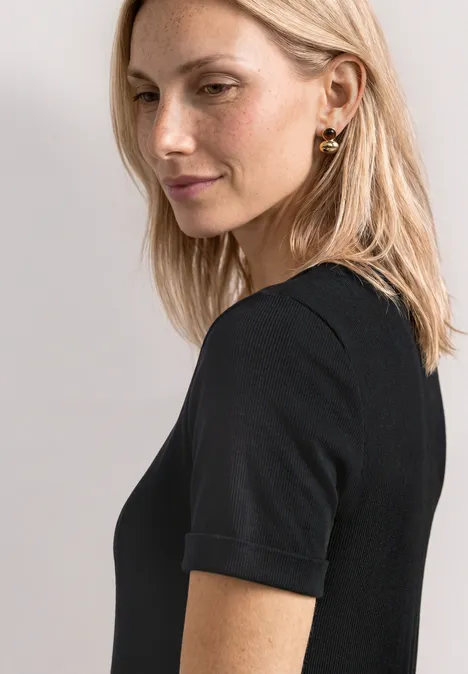Midijurk met ribbelpatroon en ronde hals Black Blonde vrouw in zwart kort T-shirt kijkt zijwaarts naar de camera, draagt gouden oorbellen.