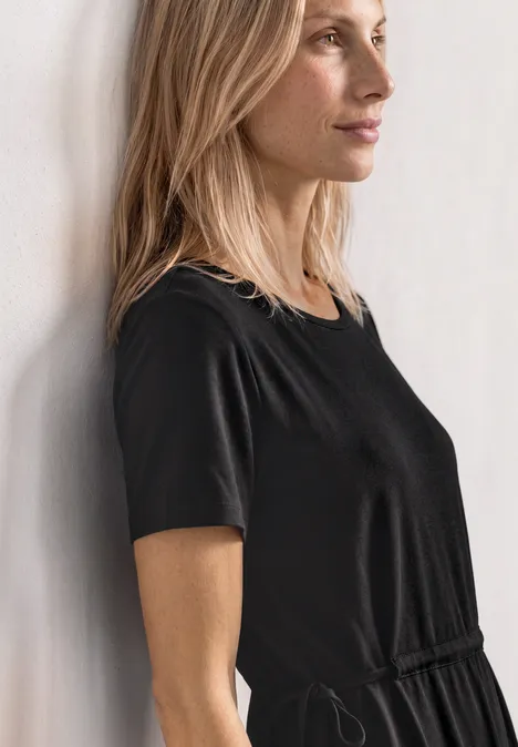 Midijurk van jersey met ronde hals Black Vrouw in zwarte jurk met korte mouwen in zijaanzicht tegen een muur geleund, kijkt richting rechts.