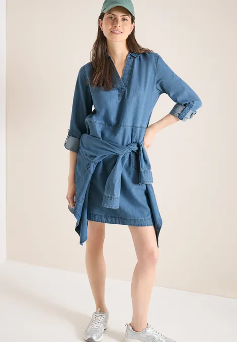 Knielange jurk in denimlook mid blue wash Jeansjurk met trui om de taille geknoopt en witte sneakers voor een lichte achtergrond.
