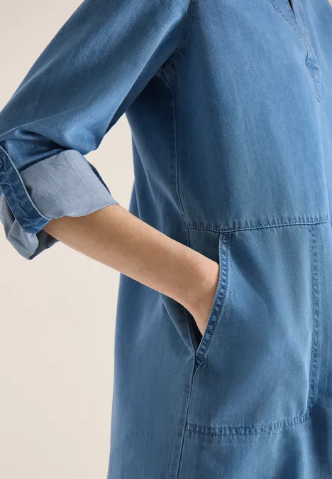 Knielange jurk in denimlook mid blue wash Persoon draagt lichtblauwe spijkerjurk met opgestroopte mouwen en heeft handen in de zijzakken gestoken.
