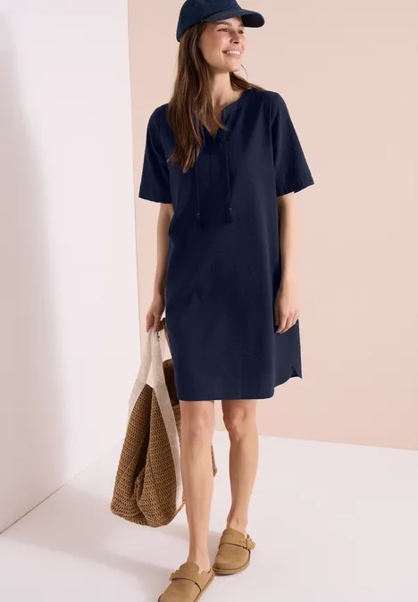 Knielange jurk in seersucker kwaliteit universal blue Vrouw in marineblauwe T-shirtjurk, pet, rieten tas en instappers tegen lichte achtergrond.