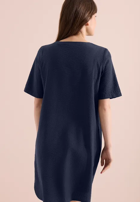 Knielange jurk in seersucker kwaliteit universal blue Een vrouw van achteren in een losvallende donkerblauwe T-shirtjurk tegen een lichte achtergrond.