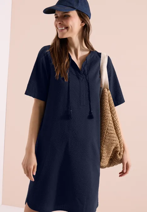 Knielange jurk in seersucker kwaliteit universal blue Vrouw draagt marineblauwe jurk met korte mouwen, V-hals en kwastjes, met pet en rieten tas.