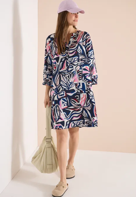 Knielange jurk met grafisch patroon universal blue Vrouw in zomerse jurk met abstract patroon, roze pet, zonnebril en schoudertas.