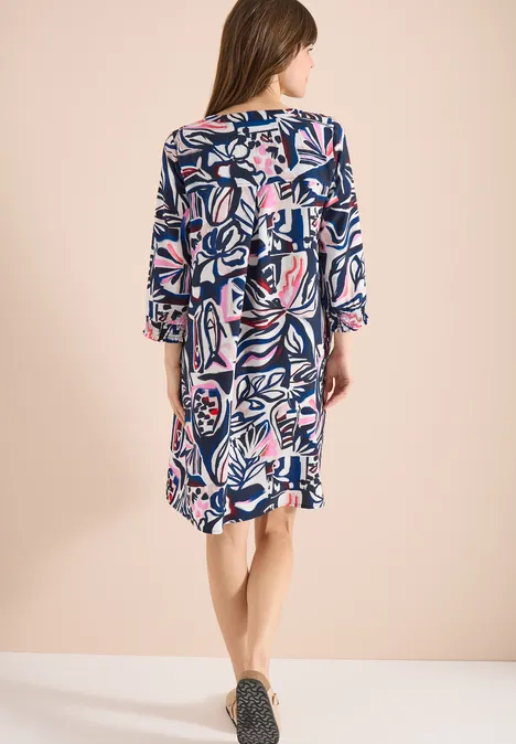 Knielange jurk met grafisch patroon universal blue Vrouw van achteren in een losse jurk met abstract blauw-roze dessin en volantmouwen voor lichte achtergrond.