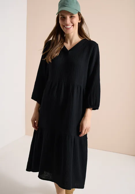 Knielange jurk in mousseline kwaliteit Black Vrouw in losse, zwarte midijurk met V-hals en driekwartmouwen voor een lichte achtergrond.