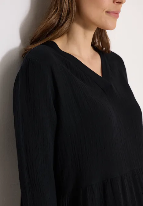 Knielange jurk in mousseline kwaliteit Black Vrouw in close-up met zwart, subtiel gestructureerd blouseje met V-hals, tegen een lichte muur.