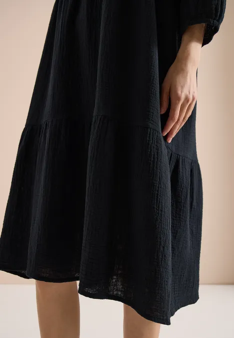 Knielange jurk in mousseline kwaliteit Black Close-up van een zwarte, getextureerde midikleding met losse pasvorm, hand zichtbaar hangend voor een lichte achtergrond.