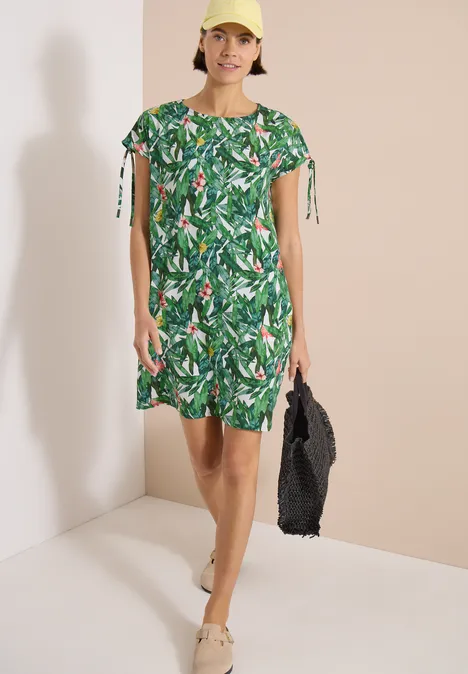 Knielange jurk met bloemenpatroon khaki Vrouw draagt groene jurk met tropisch bladpatroon, houdt een zwarte gevlochten tas en draagt beige sandalen.