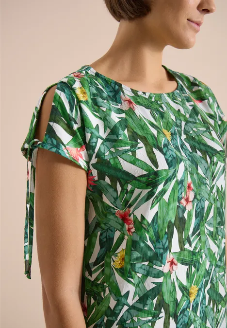 Knielange jurk met bloemenpatroon khaki Close-up van een vrouw in een groene blouse met tropische blad- en bloemenprint, mouwen met openingen en lintjes.