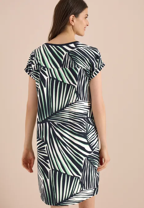 Knielange jersey jurk palm breeze green Vrouw van achteren in een jurk met korte mouwen en blauw-groene palmbladprint voor een beige achtergrond.