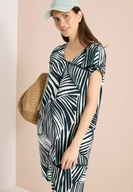 Knielange jurk met ronde V-hals universal blue Vrouw in zomerse groen-witte bladprintjurk met strooien tas voor een beige achtergrond.