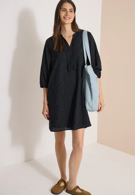 Knielange jurk met 3/4-mouwen en borduursel Black Vrouw in losvallende zwarte jurk, lichtblauwe schoudertas en bruine slippers voor een neutrale achtergrond.