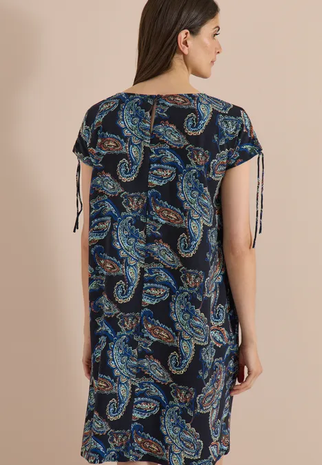 Knie-lange jurk met kapmouwtjes universal blue Vrouw van achteren in losvallende, kniehoogte jurk met blauw paisleymotief en mouwen met bindlinten.