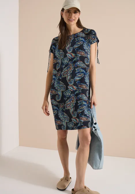 Knie-lange jurk met kapmouwtjes universal blue Vrouw in korte paisley-jurk met v-hals, pet, sandalen en een spijkerjasje in hand.