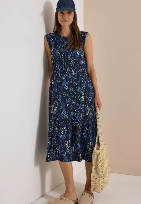 Midi-jurk met split in de hals en print urban dark blue Vrouw leunt tegen muur, draagt blauw gebloemde midijurk, witte sneakers en een strozak.