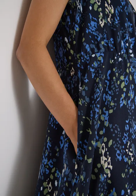 Midi-jurk met split in de hals en print urban dark blue Mouwloze donkerblauwe jurk met blauw-groene bloemenprint, arm tegen muur, hand in zak gestoken.