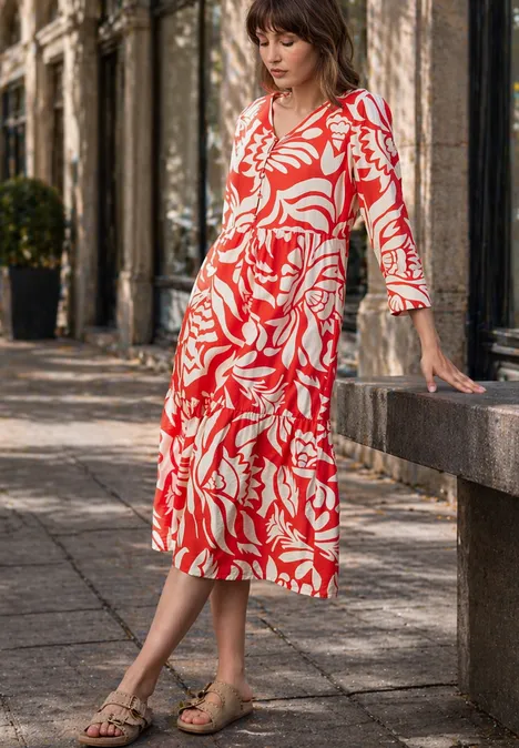 Midi-jurk met V-hals en print chili red Een vrouw in een rode midijurk met wit patroon staat buiten tegen een stenen bank.