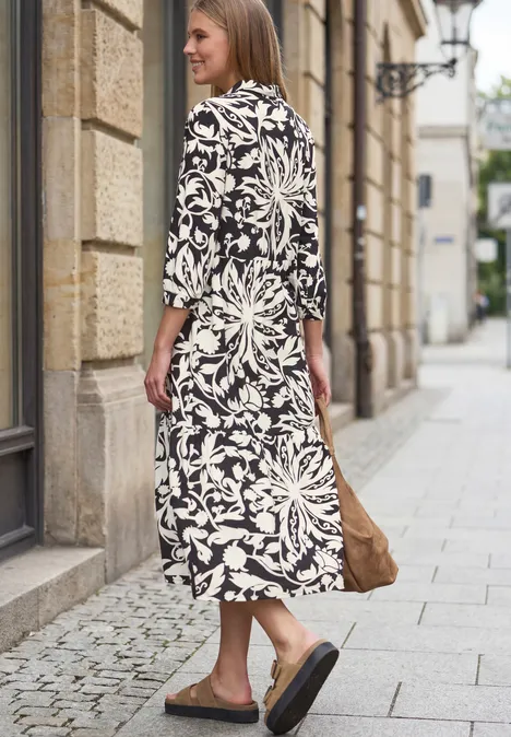 Midi-jurk met volants en bloemenpatroon Black Een vrouw in een lange zwart-witte jurk, zwarte slippers, loopt met een bruine tas langs een gevel.