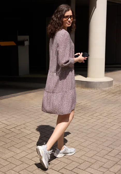Knie-lange jurk met V-hals en print vanilla sand Jonge vrouw in een gebloemde jurk en witte sneakers loopt in een zonnige stedelijke omgeving.