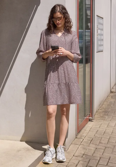 Knie-lange jurk met V-hals en print vanilla sand Vrouw in paarse jurk met stippen en witte sneakers staat bij een muur en houdt een telefoon vast.