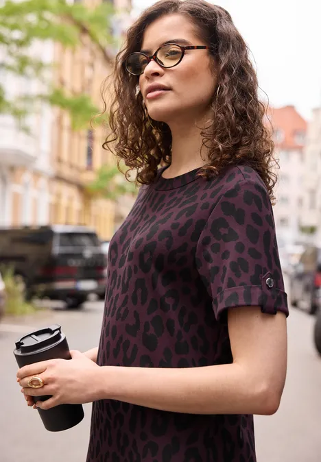 Midijurk met ronde hals en luipaardprint very berry Een vrouw met krullend haar in een luipaardprintshirt houdt een koffie to go vast op een stadsstraat.