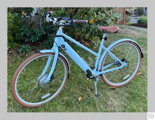 Priority eClassic Plus a blue electric bike parked on grass beside a bush