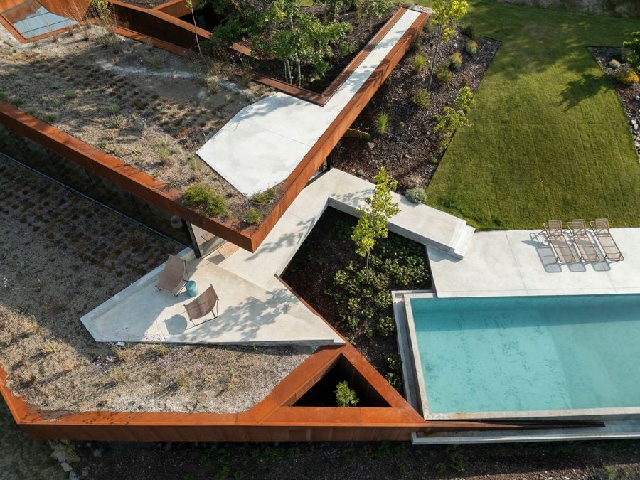 Aerial view of geometric Corten steel-framed rooftop with light concrete pathways creating angular patterns, featuring native grasses and plantings in the green roof sections, with a swimming pool visible at the edge. Aerial view of geometric Corten steel-framed rooftop with light concrete pathways creating angular patterns, featuring native grasses and plantings in the green roof sections, with a swimming pool visible at the edge.