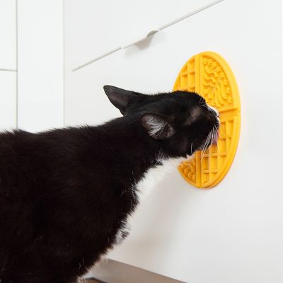 Gato negro lame una alfombrilla amarilla texturizada fijada a un armario blanco.