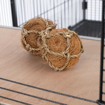 Dos pelotas de juego de materiales naturales como fibras de coco y paja trenzada, sobre una superficie de madera frente a una jaula metálica.