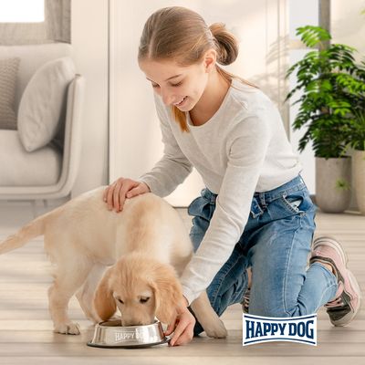 Pas jede iz zdjele s logotipom Happy Dog, u pratnji osobe. Vidljiv naziv marke: Happy Dog.