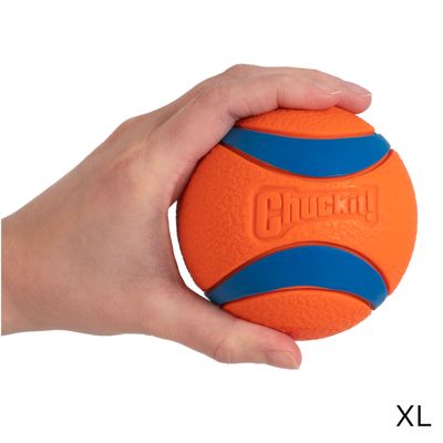 Pelota Chuckit! naranja con bandas azules, tamaño XL, sostenida por una mano. Material visible: goma.