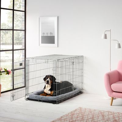 Fém kutyabox nyitott ajtóval, szürke párnával belül, modern nappaliban, rózsaszín fotellel és lámpával.