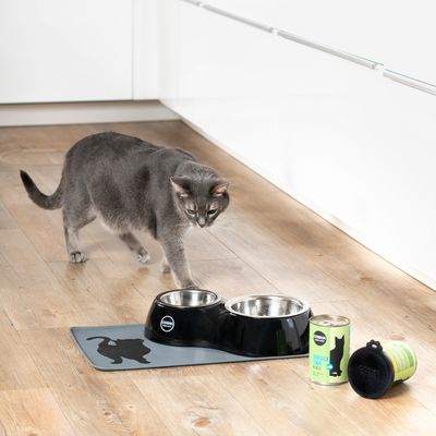 Gato gris junto a lata de comida para gatos Cosma, juego de cuencos negros con recipientes de acero inoxidable y alfombrilla a juego con motivo de gato.