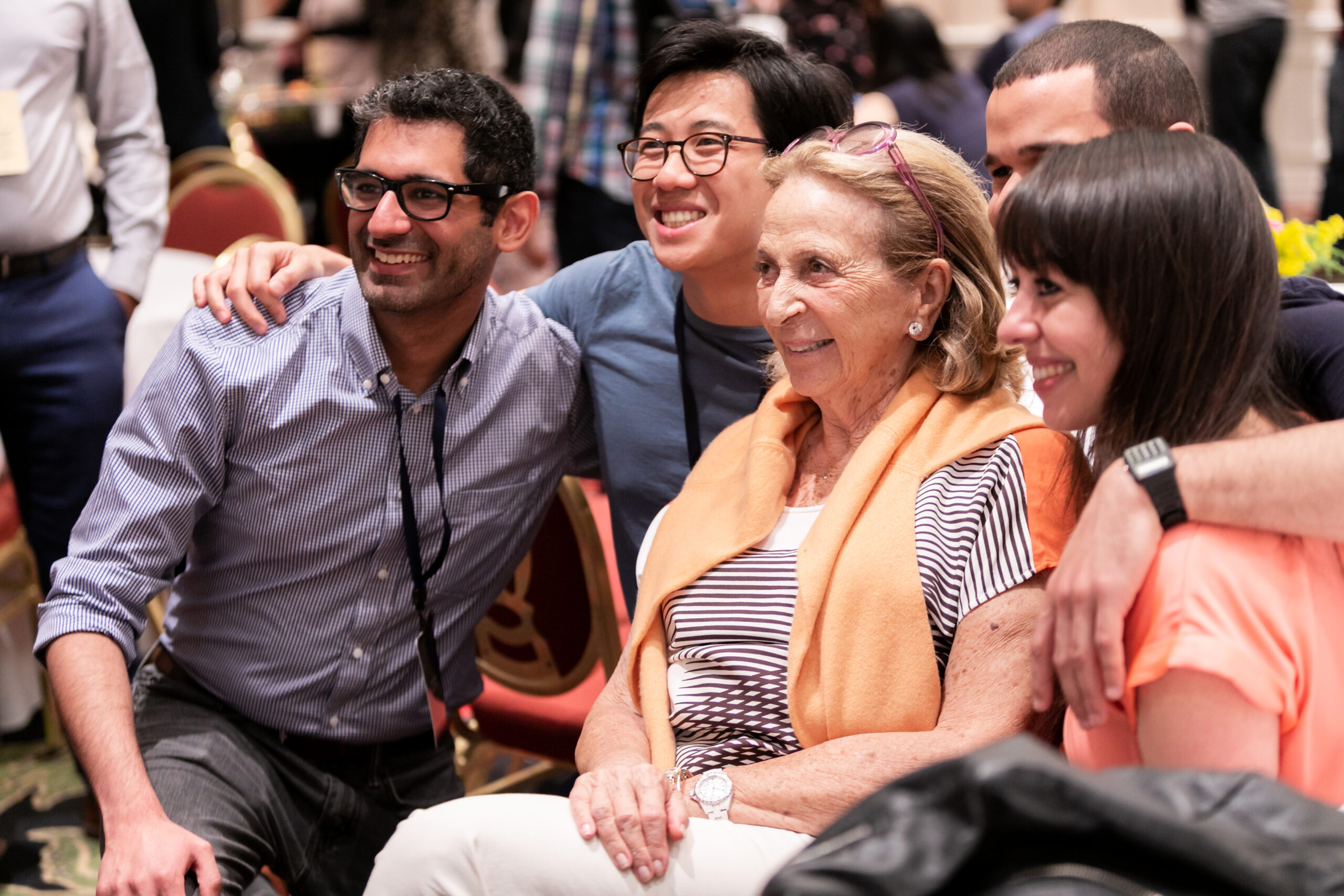Photograph of five people sitting and smiling for a photo - on a different camera. The woman in the middle is in her 80s with light skin tone and the four people around her are in their 20s of varying heritages.