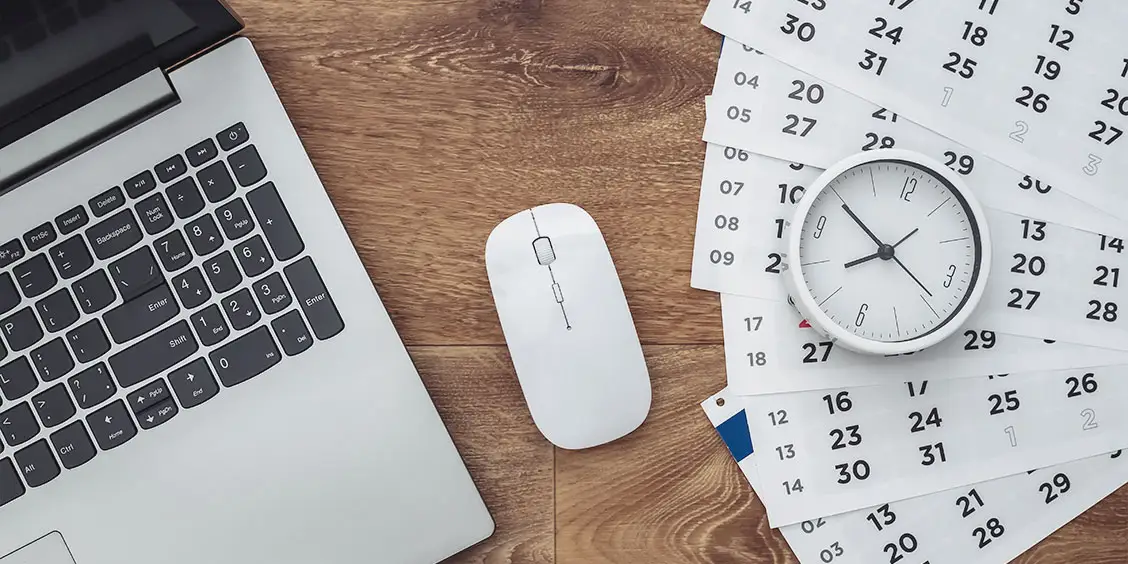 Laptop, mouse, clock, and calendar pages on a wooden desk, representing IT maintenance and time management for Piccola Tech Support Services.