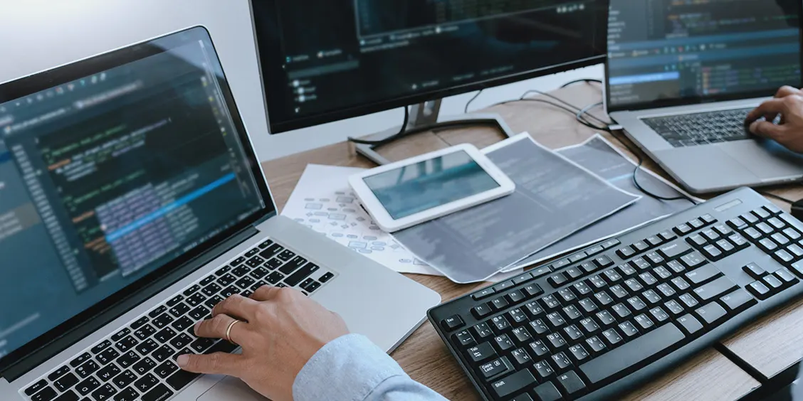 Two people at desks working on laptops with code on screens, surrounded by monitors and tech equipment in an office setting.