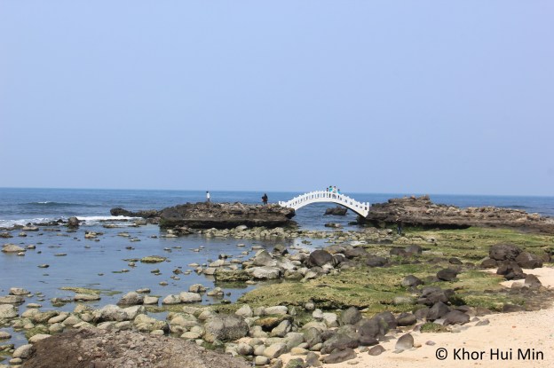 Rocky beach in front of Ximen Arch. A white Chinese bridge gleams in the sunshine. Photo taken on 12 March 2015.